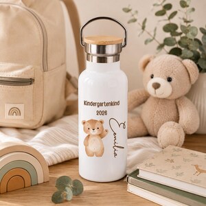 May include: A white metal water bottle with a silver handle and a light wood lid. The bottle features a brown bear illustration, the text "Kindergartenkind 2026", and the name "Emilia" printed in a dark brown script font on the front surface.