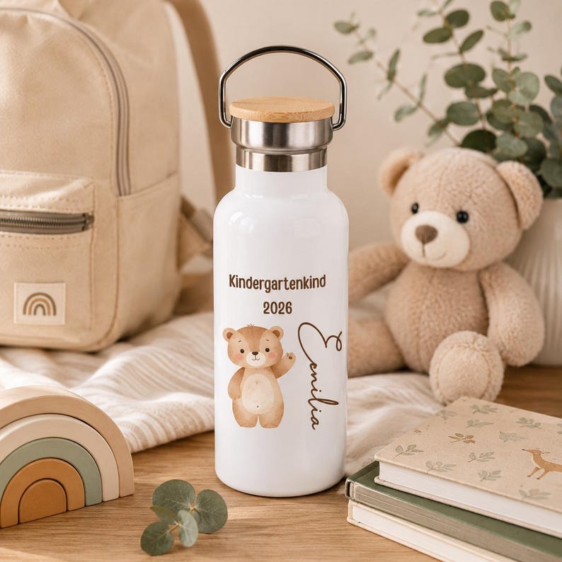 May include: A white metal water bottle with a silver handle and a light wood lid. The bottle features a brown bear illustration, the text "Kindergartenkind 2026", and the name "Emilia" printed in a dark brown script font on the front surface.
