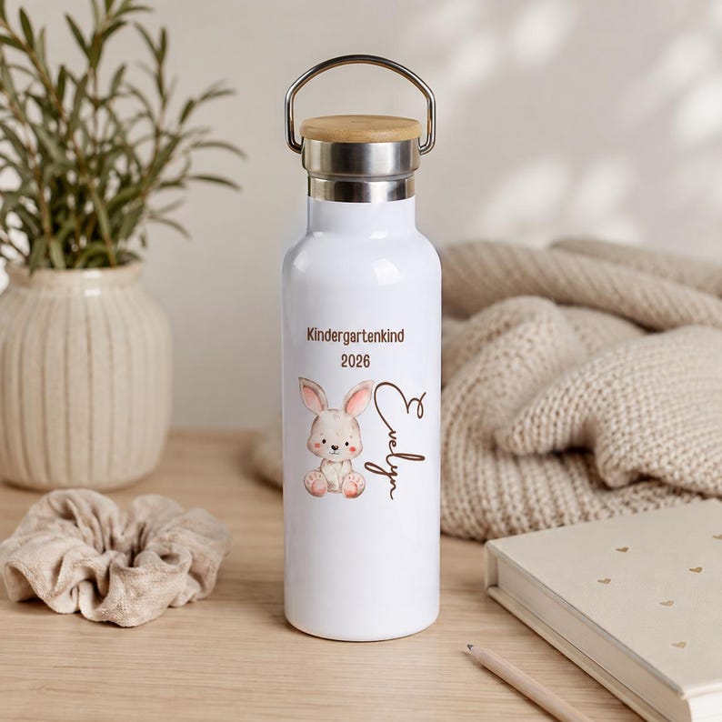 May include: A white metal water bottle with a bamboo lid and a silver handle. The bottle features a printed illustration of a small brown bunny, the text "Kindergartenkind 2026" in brown, and the name "Evelyn" written in a brown cursive script.