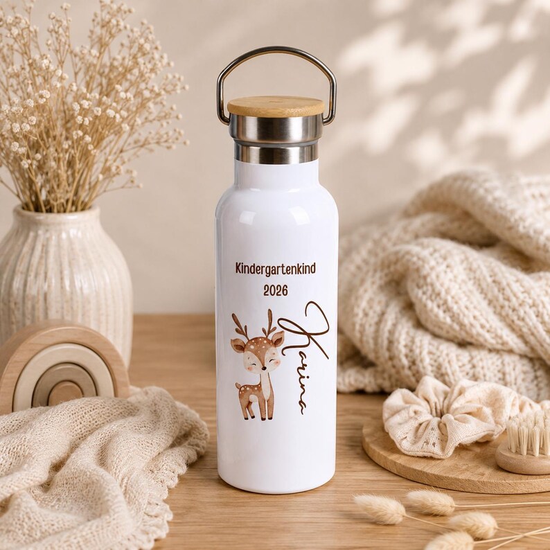 May include: A white metal water bottle with a silver rim and a wooden lid featuring a metal handle. The bottle is printed with a brown deer illustration, the text Kindergartenkind 2026, and the name Karina in a cursive brown script font.