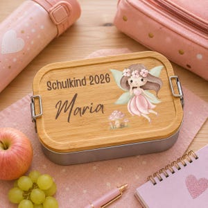 Puede incluir: Una fiambrera rectangular con tapa de bamb&uacute; que presenta una ilustraci&oacute;n impresa de un hada con cabello casta&ntilde;o, alas rosas y vestido rosa, junto a setas. El texto dice Schulkind 2026 y el nombre Maria en letra cursiva de color marr&oacute;n oscuro.
