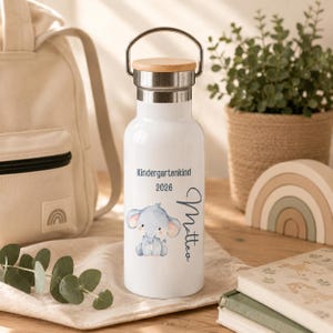 May include: A white stainless steel water bottle with a silver metal handle and a light wood lid. The bottle features a blue elephant illustration, the text Kindergartenkind 2026, and the name Matteo written in a dark blue cursive script.