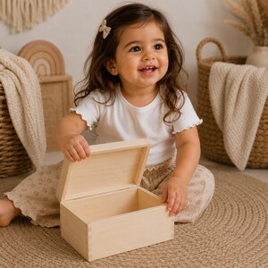 May include: A light-colored wooden box with a hinged lid, open to reveal the interior. The box is rectangular and appears to be handmade. The box is sitting on a round, woven rug. A young child is holding the box.