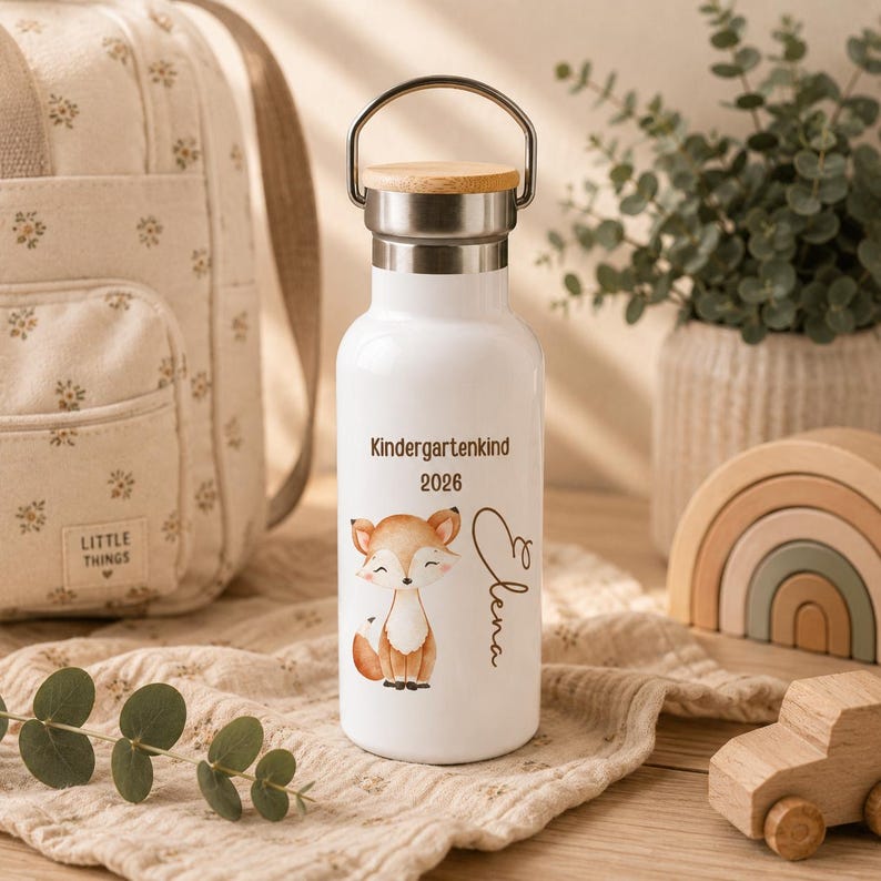 May include: A white metal water bottle with a silver neck and a wooden lid featuring a metal handle. The bottle is printed with a brown fox illustration, the text "Kindergartenkind", the year "2026", and the name "Elena" in cursive script.