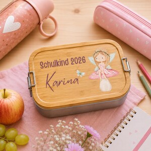 Puede incluir: Una fiambrera rectangular con tapa de bamb&uacute; que presenta una ilustraci&oacute;n impresa de un hada sentada sobre una seta, una peque&ntilde;a mariposa y el texto Schulkind 2026 y Karina. El recipiente tiene cierres de metal plateado en los laterales.