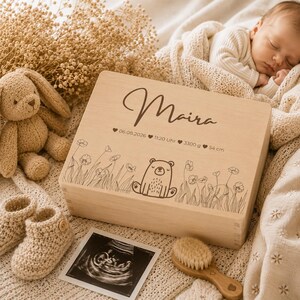May include: A light wooden keepsake box with the name "Maira" engraved on the lid, along with birth details and a bear illustration. The box is surrounded by a stuffed bunny, baby booties, an ultrasound image, and a baby brush.