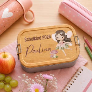 Puede incluir: Una fiambrera rectangular con tapa de bamb&uacute; claro que presenta una ilustraci&oacute;n impresa de un hada de pelo oscuro, una seta y el texto "Schulkind 2026" y "Paulina" en fuentes de color marr&oacute;n oscuro con cierres met&aacute;licos laterales.