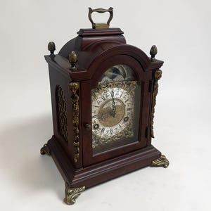 Könnte beinhalten: Eine dunkelbraune Kaminuhr aus Holz mit Messinggriff und dekorativen Akzenten. Die Uhr hat eine Glasfront, ein detailliertes Zifferblatt und Messingfüße. Die Uhr ist ein dekorativer Artikel im Vintage-Stil.