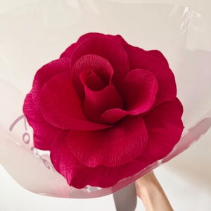 May include: A large, vibrant red paper rose, meticulously crafted with textured petals. The oversized flower is wrapped in clear cellophane, held by a gray ribbon. The rose is a striking decorative piece.