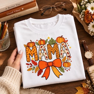 May include: A white t-shirt featuring the word MAMA in large, patterned block letters. The design includes orange pumpkins, colorful autumn leaves, and a large orange bow at the bottom. The letters have a leopard print and leaf pattern.