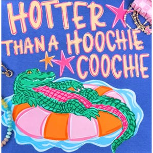 Może przedstawiać: Niebieska grafika z napisem "HOTTER THAN A HOOCHIE COOCHIE" w kolorze różowym i żółtym. Zielony aligator unosi się na różowo-pomarańczowym kole do pływania. Akcesoria, takie jak bransoletka i naszyjnik, są w tle.