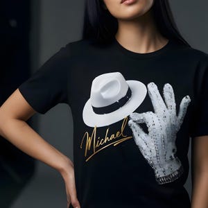 Puede incluir: Camiseta negra con un sombrero fedora blanco, un guante con lentejuelas y el nombre "Michael" en oro. El sombrero tiene una banda negra y el guante está detallado con lentejuelas plateadas. La camiseta tiene cuello redondo.