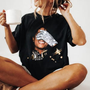 May include: Black t-shirt featuring a graphic of a smiling person with a sparkling glove covering one eye. The shirt has gold and silver embellishments. A white mug is held in the person's hand.