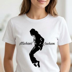 Könnte beinhalten: Weißes T-Shirt mit einer schwarzen Silhouette einer Person in einer Tanzpose, mit dem Namen "Michael Jackson" in Schreibschrift. Das Design befindet sich auf der Vorderseite. Das T-Shirt hat kurze Ärmel und einen Rundhalsausschnitt.