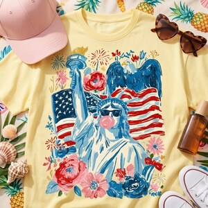 Könnte beinhalten: Gelbes T-Shirt mit einer Grafik der Freiheitsstatue mit Sonnenbrille und Kaugummiblase. Das Design umfasst eine amerikanische Flagge, einen Adler und Blumen. Eine rosa Baseballkappe und herzförmige Sonnenbrillen sind ebenfalls zu sehen.