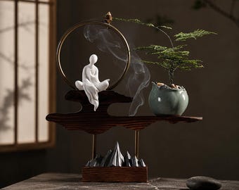 Op de afbeelding: Een decoratieve wierookbrander met een wit keramieken figuur op een donkerhouten voet, een gouden metalen ring en een kleine groene plant in een lichtblauwe keramieken pot. Rookpluimen stijgen op van een wierookkegel boven op de gouden ring.