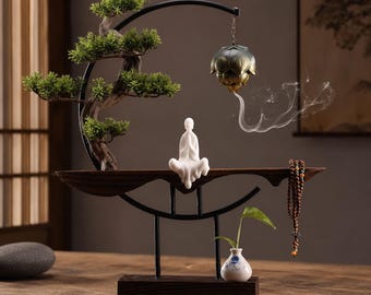 Op de afbeelding: Een decoratief display met een wit keramieken mediterend figuur, een groene kunstmatige bonsaiboom en een hangende metalen lotus wierookbrander die rook afgeeft. Een kleine witte vaas met een groen blaadje en houten gebedskralen zijn inbegrepen.