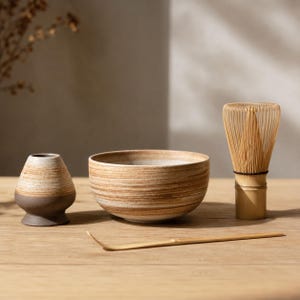 May include: A matcha tea set featuring a ceramic bowl, a whisk, a whisk holder, and a bamboo scoop. The bowl and holder have a brown and beige striped design. The whisk is made of bamboo.