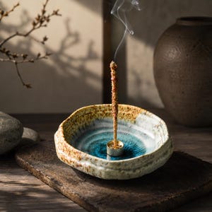 May include: A round, shallow ceramic incense burner with a textured, speckled beige exterior and a vibrant blue, crackled glaze interior. A small, circular brass incense holder sits in the centre, holding a single burning incense stick with smoke.