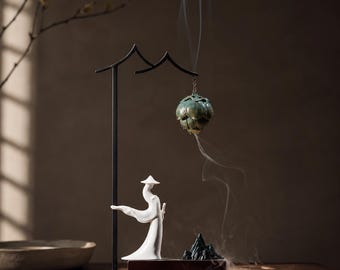 Op de afbeelding: Een zwarte metalen standaard houdt een hangende groene lotusvormige wierookbrander vast die rook uitstoot. Daaronder staat een wit keramieken figuur met een conische hoed op een rechthoekige houten voet naast een kleine, donkere wierookhouder.