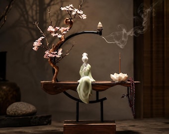 Op de afbeelding: Een decoratieve wierookhouder met een serene porseleinen beeldje in een vloeiende groene jurk. De houder heeft een houten basis, een gebogen zwarte metalen arm en een kleine kersenbloesemboom. Wierookrook stijgt op.