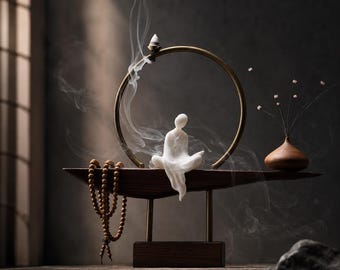 Op de afbeelding: Een zen-geïnspireerde wierookhouder met een mediterende figuur, een houten dienblad en een goudkleurige ring. Een snoer van houten kralen hangt aan de linkerkant. Een kleine vaas met delicate stelen staat rechts. Wispeltjes rook stijgen op van de wierookkegel.