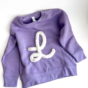 May include: A lavender sweatshirt with a white, fluffy letter "L" appliqued on the front. The long-sleeved top has a crew neck and ribbed cuffs and hem. The garment is made of a soft material.