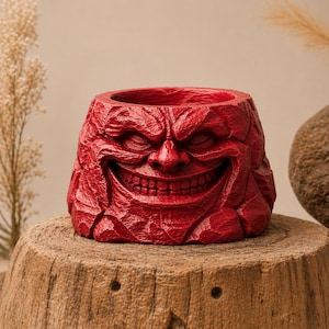 May include: A red, textured planter with a carved face design. The planter has a wide opening and a smiling face carved into the front. The planter is sitting on a wooden surface.