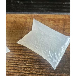 Puede incluir: Un paquete de plástico transparente y sellado, posiblemente conteniendo un líquido o gel, descansa sobre una superficie de madera. El paquete tiene forma de almohada con arrugas visibles. El fondo es una superficie oscura y texturizada.