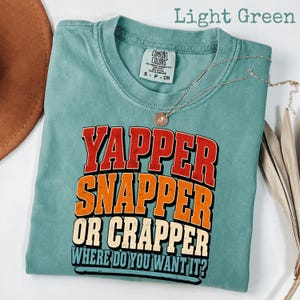 Può includere: Una t-shirt a maniche corte color verde chiaro con il testo YAPPER SNAPPER OR CRAPPER WHERE DO YOU WANT IT in lettere stampatello rosse, arancioni e color crema. La maglia presenta un'etichetta Comfort Colors sul colletto e una catenina.