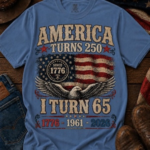 Könnte beinhalten: Hellblaues T-Shirt mit patriotischem Design. Das Shirt zeigt den Text "AMERICA TURNS 250" über einer amerikanischen Flagge und einem Adler. Darunter steht "I TURN 65" mit den Jahren 1776-1961-2026.