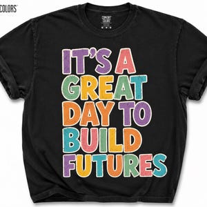 May include: Black Comfort Colors t-shirt with the text "IT'S A GREAT DAY TO BUILD FUTURES" in colorful, outlined letters. The shirt has a crew neck and short sleeves, perfect for casual wear.
