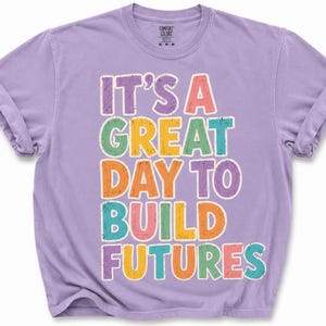 May include: A lilac-colored t-shirt with rolled-up sleeves. The shirt features the text "IT'S A GREAT DAY TO BUILD FUTURES" in colorful, block letters. The shirt is made of a soft, comfortable material.