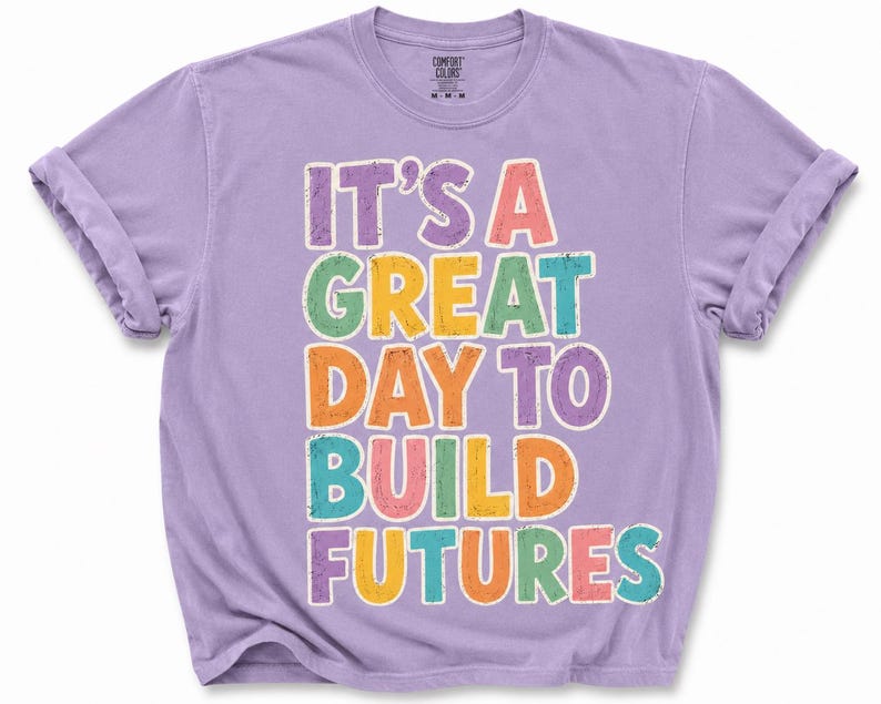 May include: A lilac-colored t-shirt with rolled-up sleeves. The shirt features the text "IT'S A GREAT DAY TO BUILD FUTURES" in colorful, block letters. The shirt is made of a soft, comfortable material.