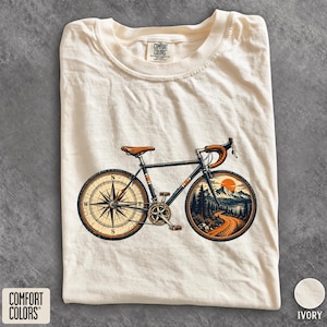 Può includere: Una t-shirt a maniche corte color avorio con una stampa grafica di una bicicletta. La ruota posteriore presenta un design a bussola, mentre quella anteriore mostra un paesaggio montano con una strada tortuosa e un sole. Etichetta Comfort Colors.
