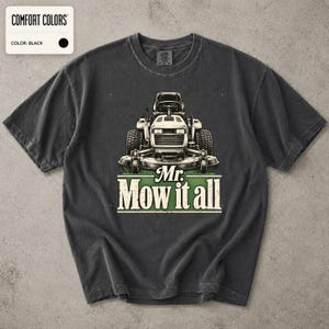 Pode incluir: Uma t-shirt preta de manga curta com um gráfico de um trator corta-relvas centrado no peito. Abaixo do corta-relvas, o texto "Mr. Mow it all" está impresso numa fonte clara e em negrito sobre um elemento de fundo retangular verde.