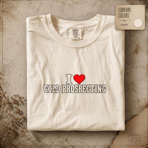Peut inclure: Un t-shirt à manches courtes de couleur ivoire avec le texte I [symbole cœur] GOLD PROSPECTING imprimé en noir avec un cœur rouge. Le t-shirt présente une encolure ronde et une étiquette de marque Comfort Colors indiquant une taille medium.