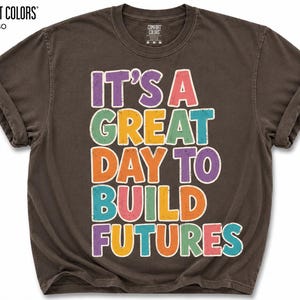 May include: Espresso-colored t-shirt with rolled sleeves. The shirt features the text "IT'S A GREAT DAY TO BUILD FUTURES" in colorful, outlined letters. The brand name "COMFORT COLORS" is printed at the top left.