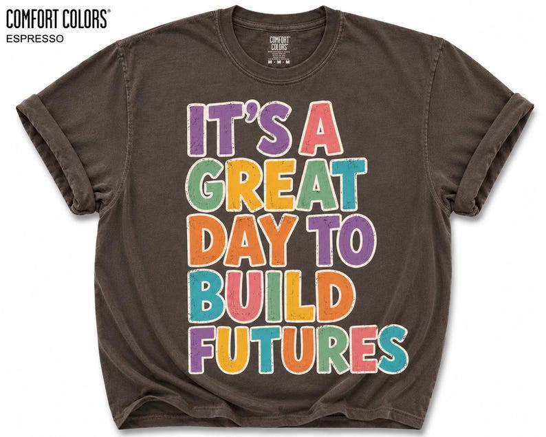 May include: Espresso-colored t-shirt with rolled sleeves. The shirt features the text "IT'S A GREAT DAY TO BUILD FUTURES" in colorful, outlined letters. The brand name "COMFORT COLORS" is printed at the top left.