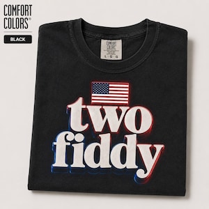 Può includere: Una t-shirt nera a maniche corte con la scritta 'two fiddy' in lettere bianche con ombreggiature rosse e blu. Sopra il testo è presente una piccola grafica della bandiera americana. La maglietta presenta un'etichetta del marchio Comfort Colors.