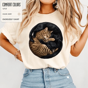 Puede incluir: Una camiseta oversize de color marfil con un gráfico circular de un gato atigrado marrón y un gato negro enroscados en forma de yin-yang. La camiseta Comfort Colors 1717 tiene mangas cortas y cuello redondo con una ilustración detallada.