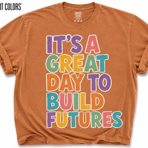 May include: A yam-colored t-shirt with rolled-up sleeves. The shirt features the text "IT'S A GREAT DAY TO BUILD FUTURES" in colorful, outlined letters. The shirt is from Comfort Colors.