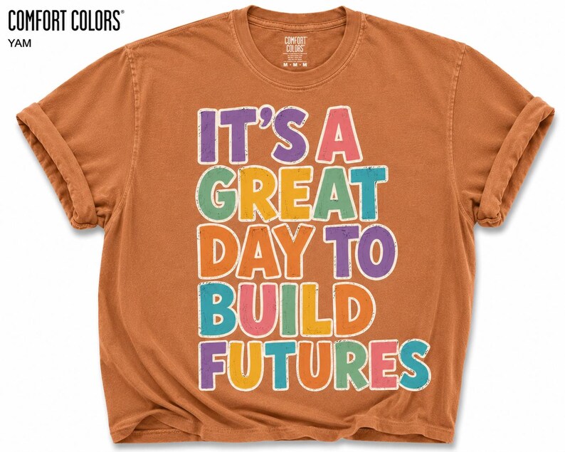 May include: A yam-colored t-shirt with rolled-up sleeves. The shirt features the text "IT'S A GREAT DAY TO BUILD FUTURES" in colorful, outlined letters. The shirt is from Comfort Colors.