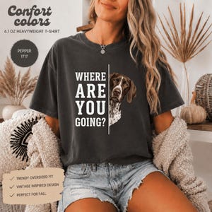 Può includere: Una t-shirt grigio antracite pesante con la grafica di un cane marrone e bianco che sbuca da dietro una linea verticale. Il testo "WHERE ARE YOU GOING?" è stampato in lettere maiuscole bianche. La t-shirt ha un girocollo e maniche corte.