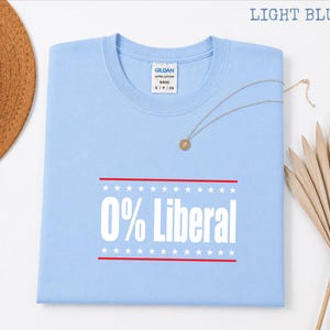 May include: Light blue t-shirt with the text "0% Liberal" in white, framed by red lines and stars. The shirt has a crew neck and a Gildan label. A gold necklace and a straw hat are also visible.