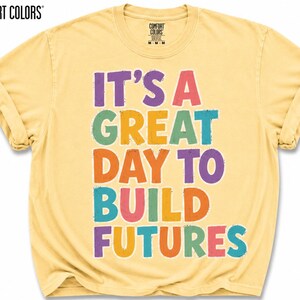 May include: Butter-colored t-shirt with rolled sleeves. The front features the text "IT'S A GREAT DAY TO BUILD FUTURES" in colorful, bold letters. The Comfort Colors brand logo is visible at the top left.