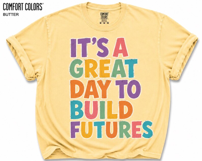 May include: Butter-colored t-shirt with rolled sleeves. The front features the text "IT'S A GREAT DAY TO BUILD FUTURES" in colorful, bold letters. The Comfort Colors brand logo is visible at the top left.