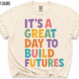 May include: Ivory-colored Comfort Colors t-shirt with the text "IT'S A GREAT DAY TO BUILD FUTURES" in colorful, block letters. The shirt has rolled-up sleeves and a crew neck. The Comfort Colors logo is visible at the top.