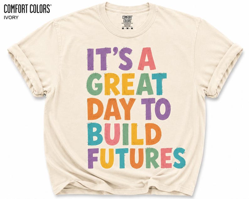 May include: Ivory-colored Comfort Colors t-shirt with the text "IT'S A GREAT DAY TO BUILD FUTURES" in colorful, block letters. The shirt has rolled-up sleeves and a crew neck. The Comfort Colors logo is visible at the top.