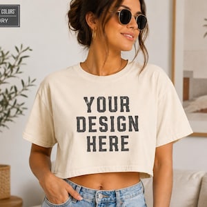 Könnte beinhalten: Ein elfenbeinfarbenes, kurzärmeliges bauchfreies T-Shirt mit dem schwarzen Schriftzug "YOUR DESIGN HERE", der in einer fetten Blockschrift mittig auf der Brust aufgedruckt ist. Das Kleidungsstück hat einen Rundhalsausschnitt und einen lockeren Schnitt.